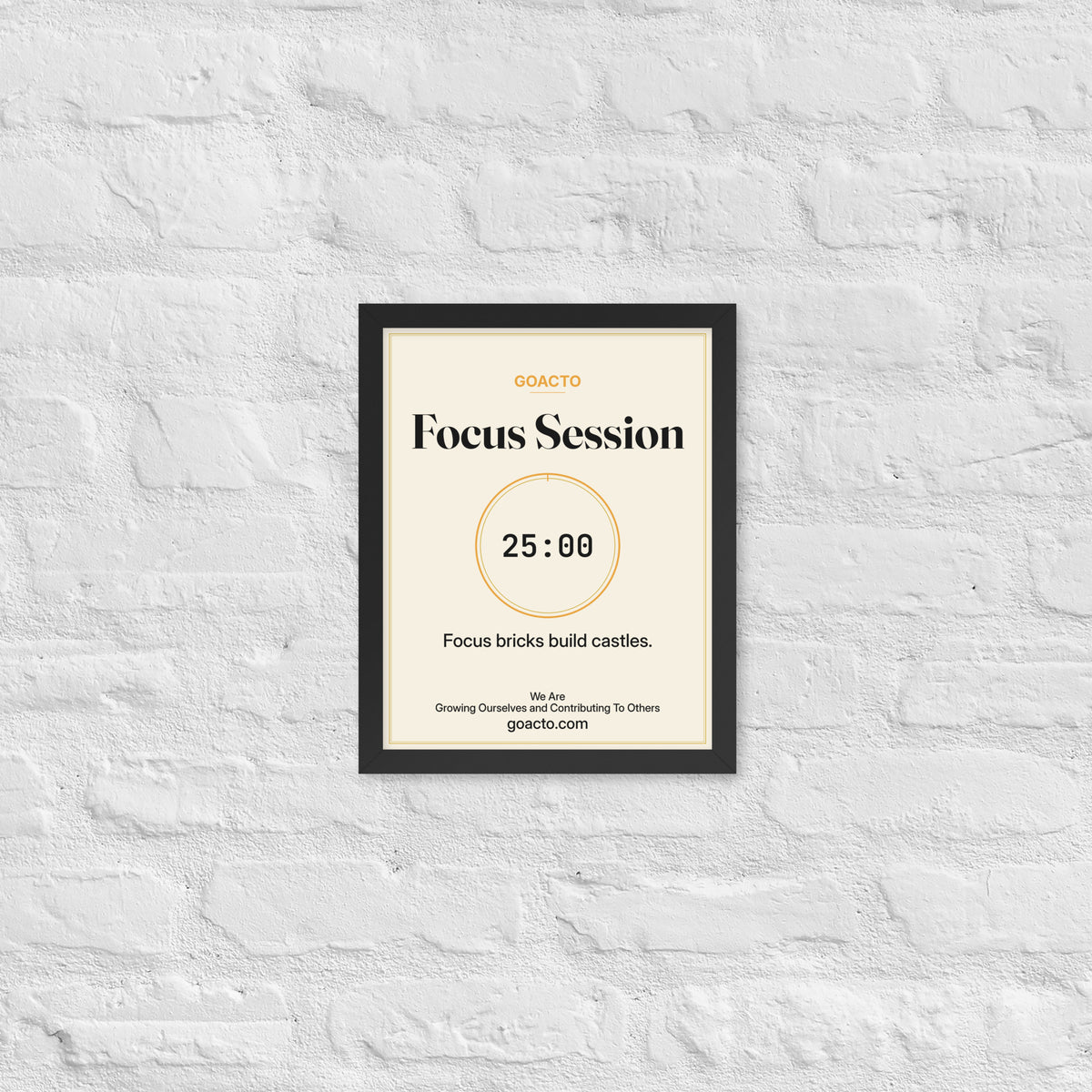 Focus Session Framed Print, Cream design, 11x14 in a black frame on a white brick wall: thin amber ring around a dark 25:00 timer, with dark type on a cream background, the line 'Focus bricks build castles.' and 'We Are / Growing Ourselves and Contributing To Others' above goacto.com
