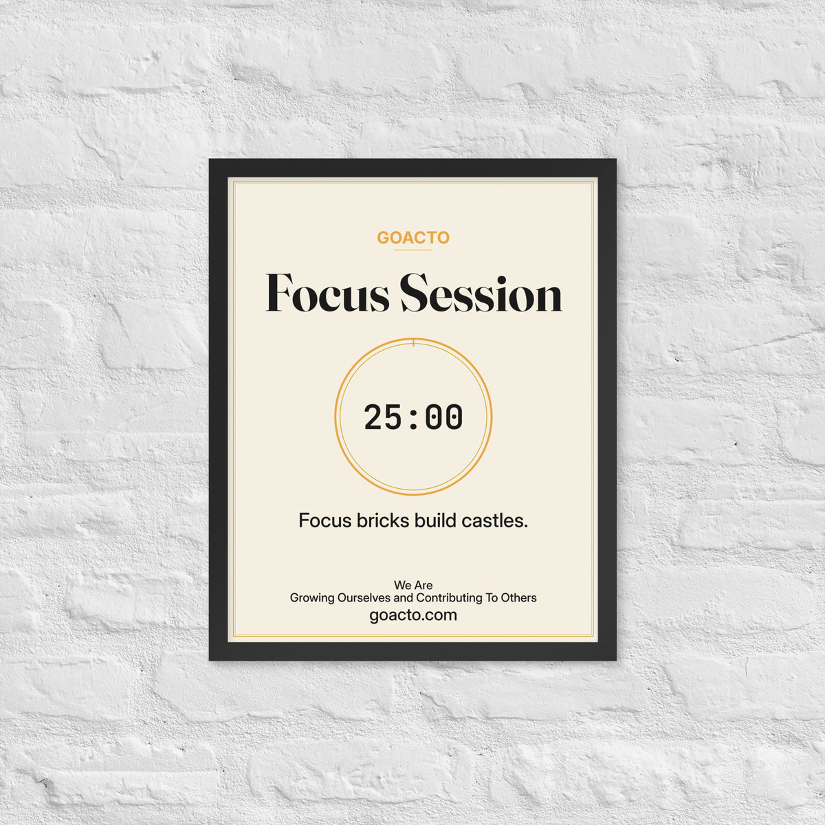 Focus Session Framed Print, Cream design, 16x20 in a black frame on a white brick wall: thin amber ring around a dark 25:00 timer, with dark type on a cream background, the line 'Focus bricks build castles.' and 'We Are / Growing Ourselves and Contributing To Others' above goacto.com