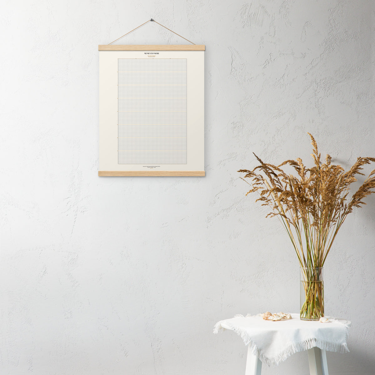 Memento Mori Tracker by GOACTO (16×20, Oak Hanger) mockup: cream week grid poster hung from an oak wood hanger on a white textured wall, beside a vase of dried grasses